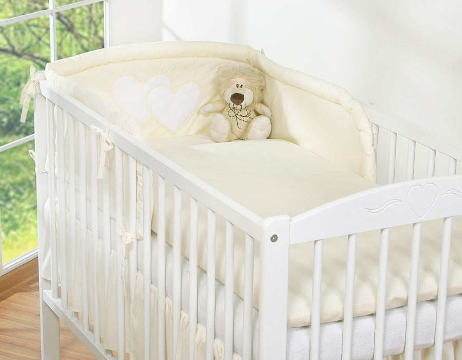White crib with beige bedding and a teddy bear in a bright room.