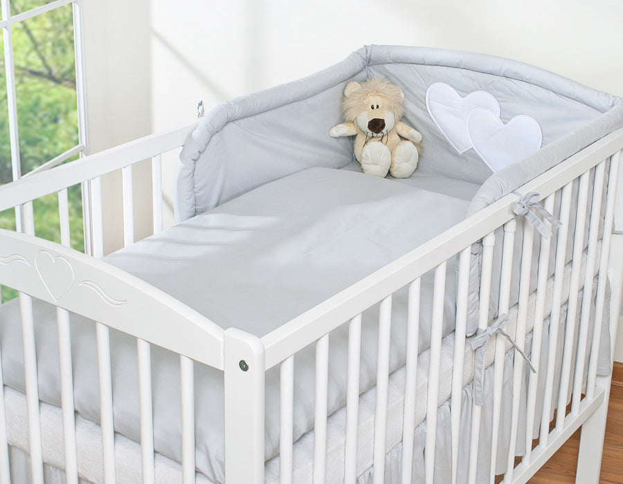 White crib with gray bedding and a teddy bear, set against a blurred indoor background.