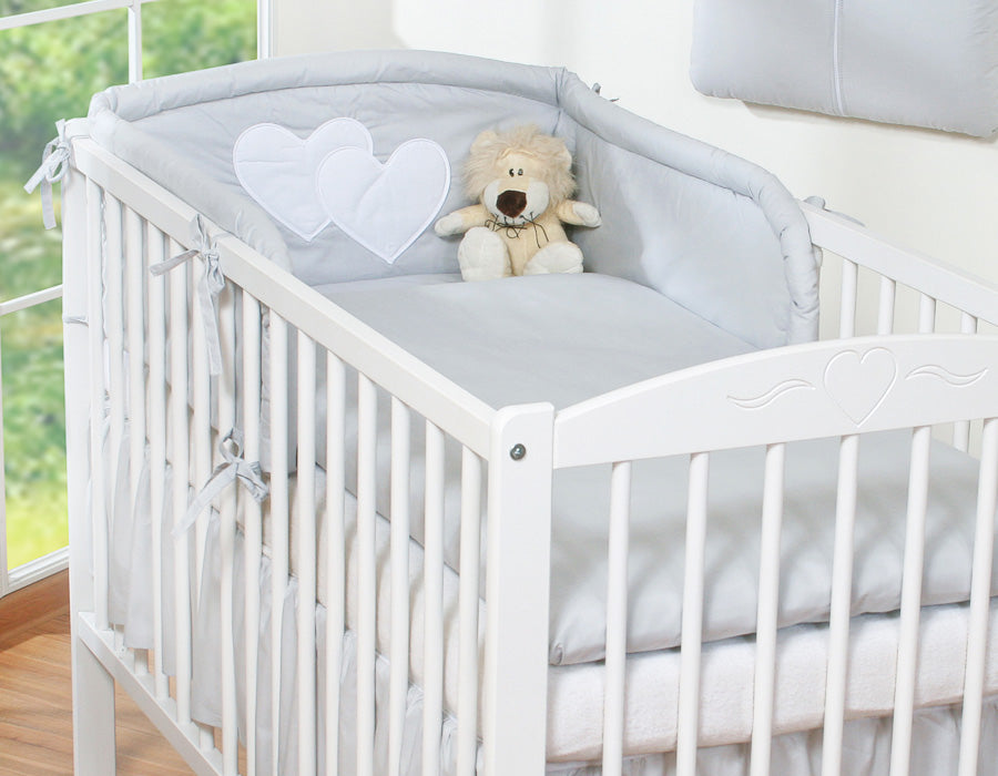 White crib with gray bedding, heart-shaped pillow, and teddy bear in a bright room.