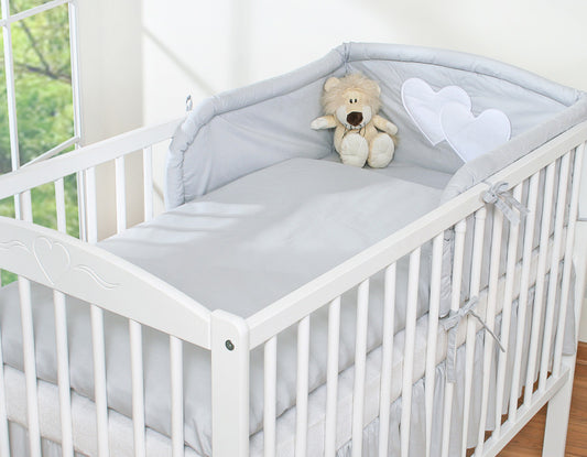 White crib with gray bedding and a teddy bear, set against a blurred indoor background.