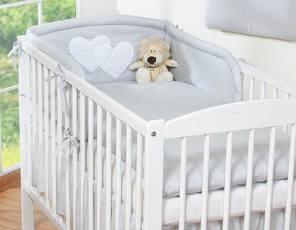 White crib with gray bedding, heart-shaped pillow, and teddy bear in a bright room.
