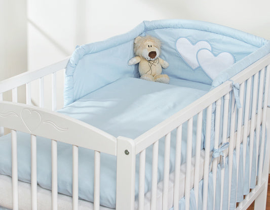 White crib with blue bedding and a teddy bear, set against a white wall.